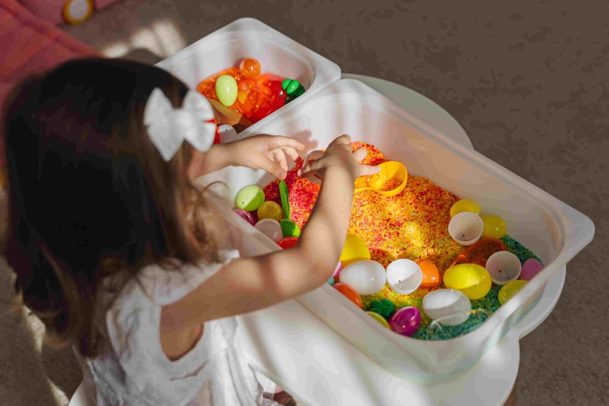 Girl playing in a sensory bin filled with colored rice and eggs. 