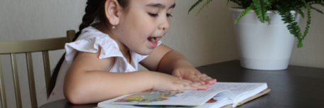 Young girl reading excitedly from a book while sitting at the kitchen table. 