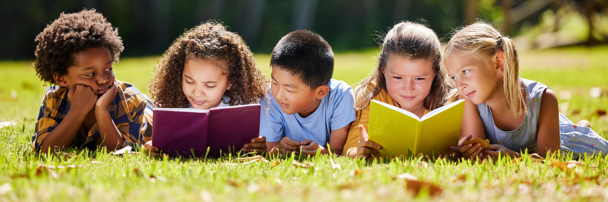 A group of kindergarteners reading books on a grassy lawn. 