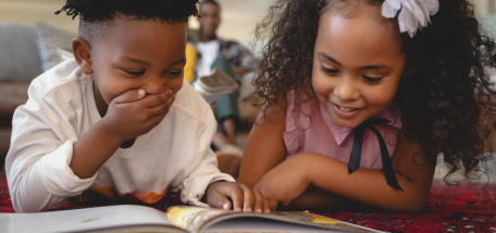 Two small children looking at a book together and laughing. 