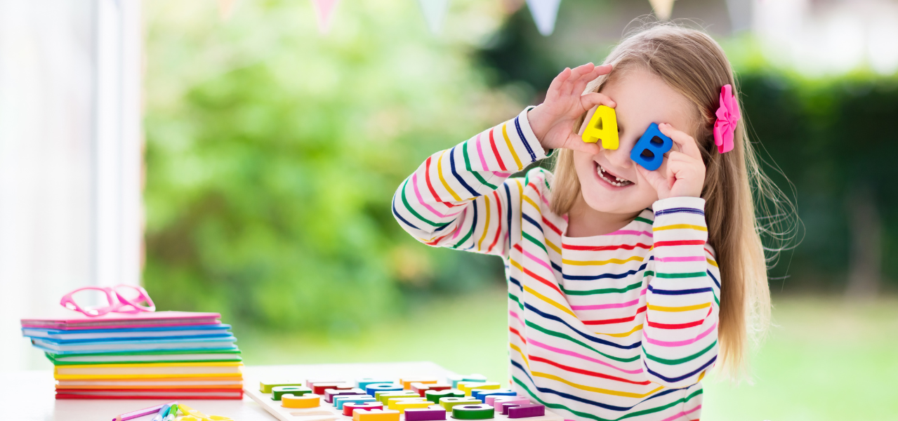 Kindergarten girl holding a yellow letter A and a blue letter b up to her eyes while making a silly face. 