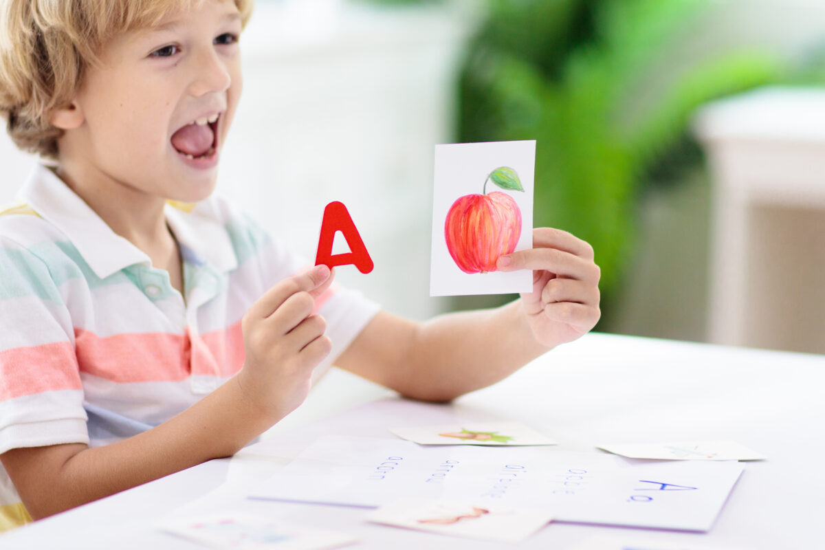 Little boy holding flashcards of the letter a and an apple making the letter a sound with his mouth. 