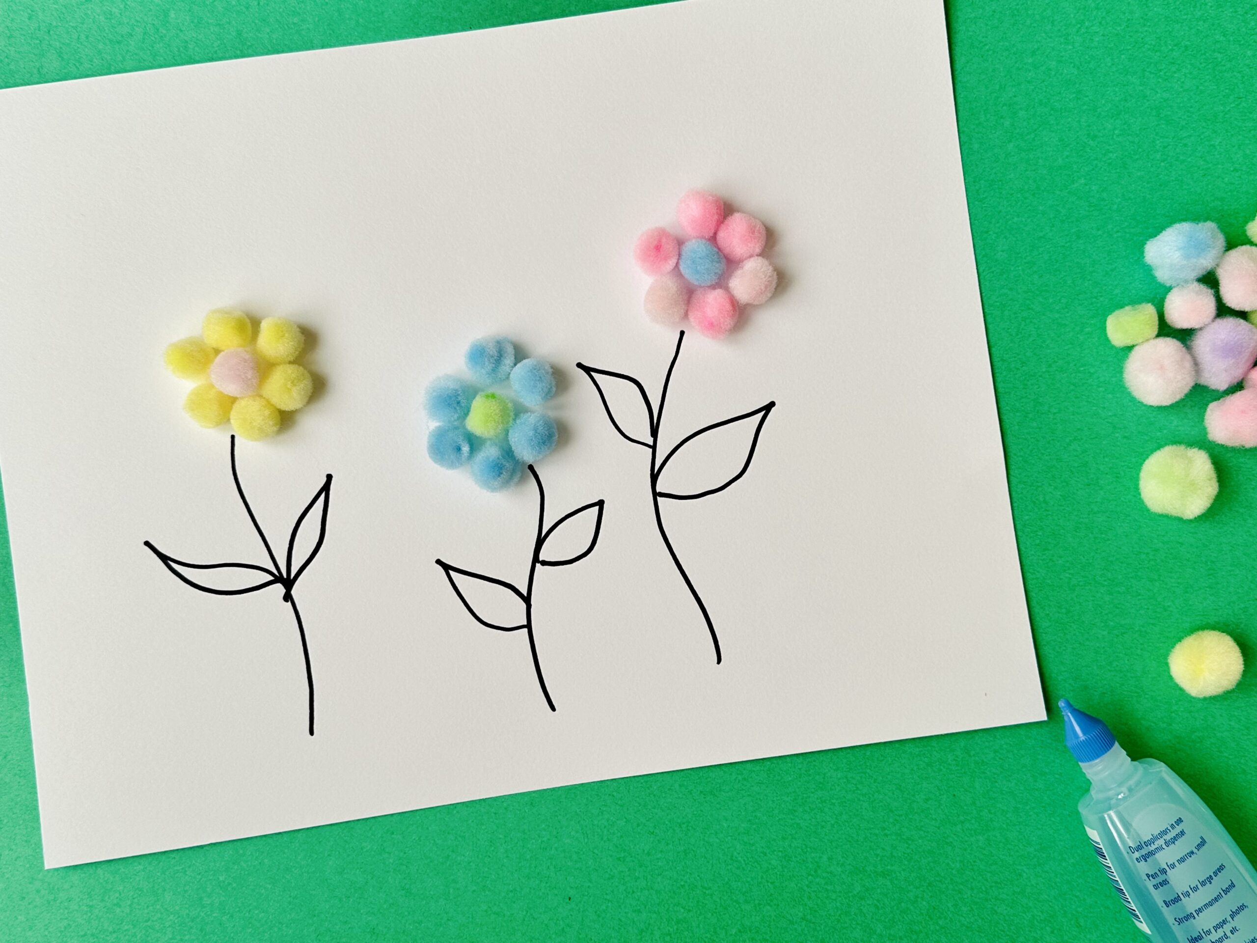 Drawn flower stems topped with colorful pom-poms as blossoms, with glue bottle and extra pom-poms nearby.