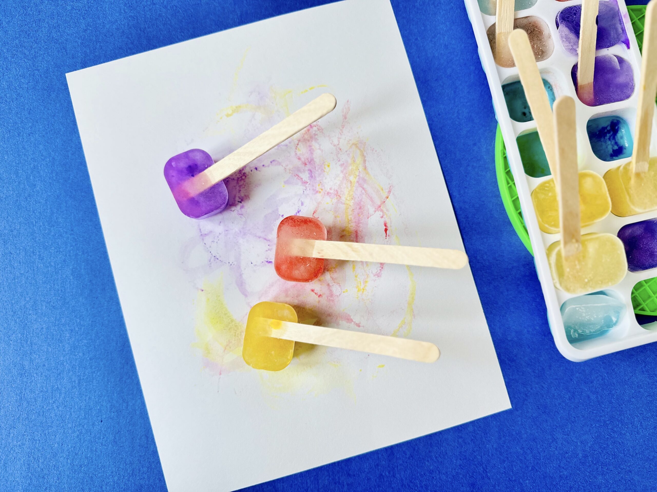 Colorful melting ice paint cubes with craft stick handles being used to paint on white paper.