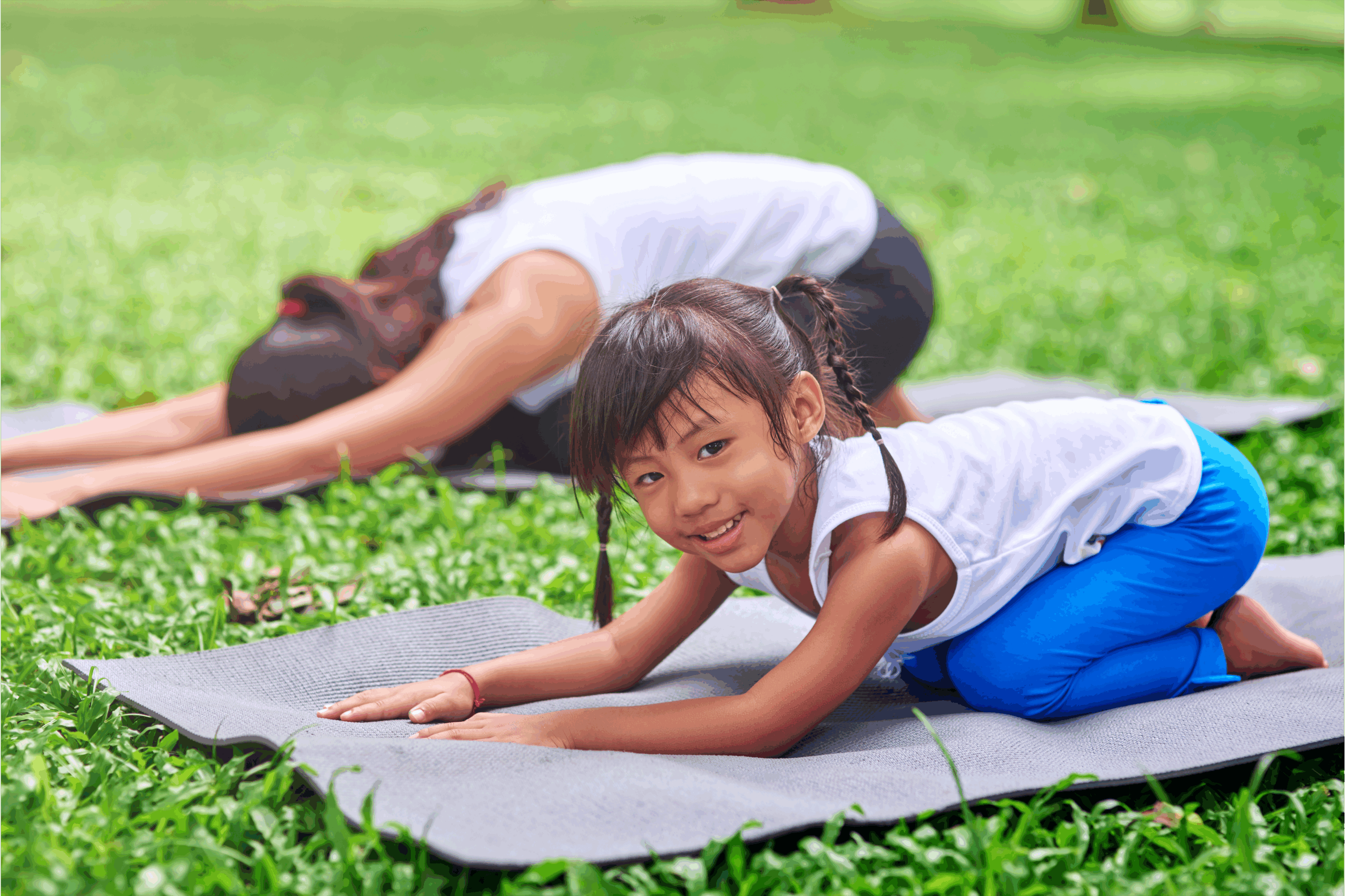 A young girl and her mother doing yoga on a mat outside. 