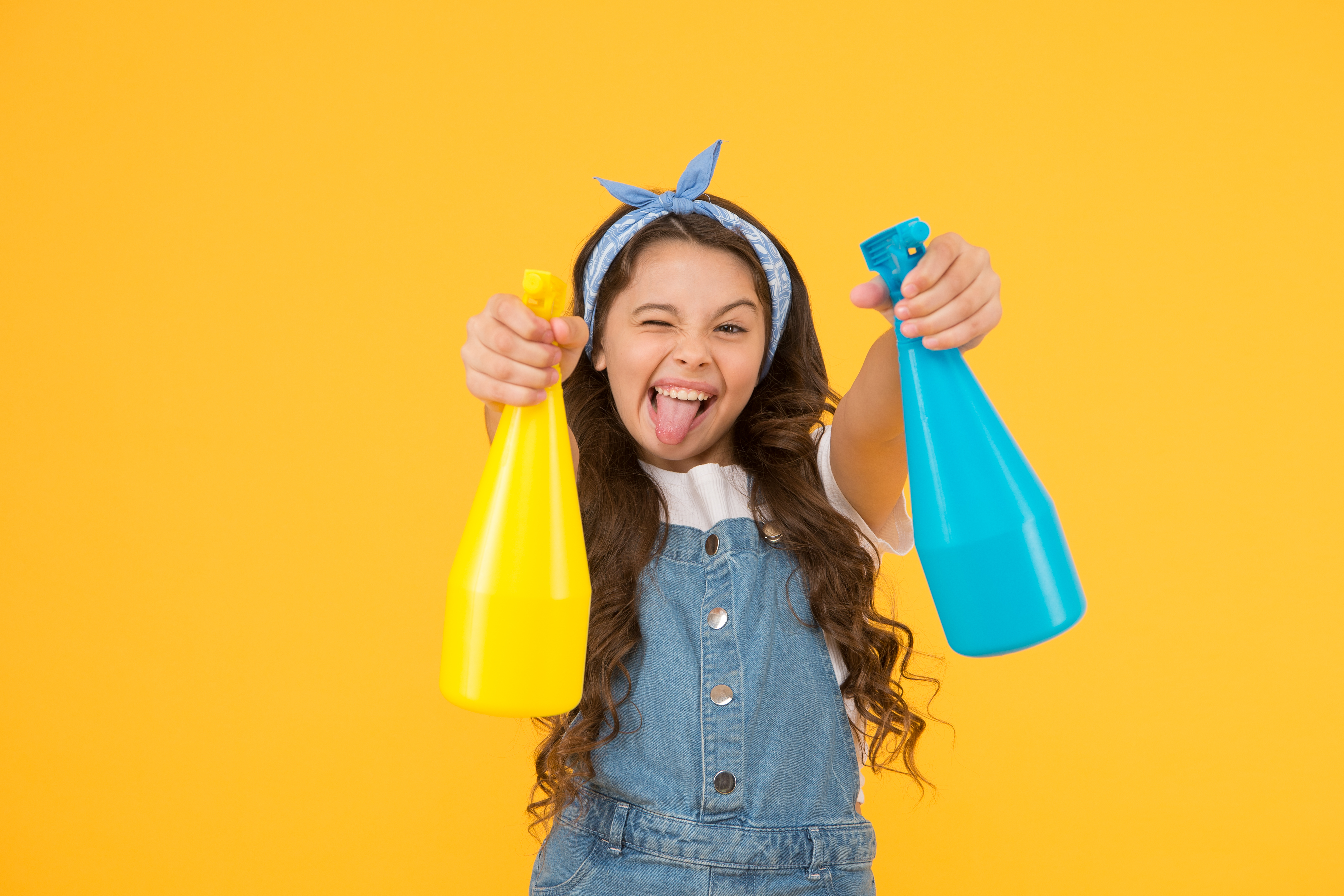 A young girl holding colorful spray bottles in front of a yellow backdrop. 