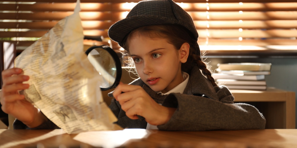 A young girl dressed up in a detective suit using a magnifying glass to look at a paper. 