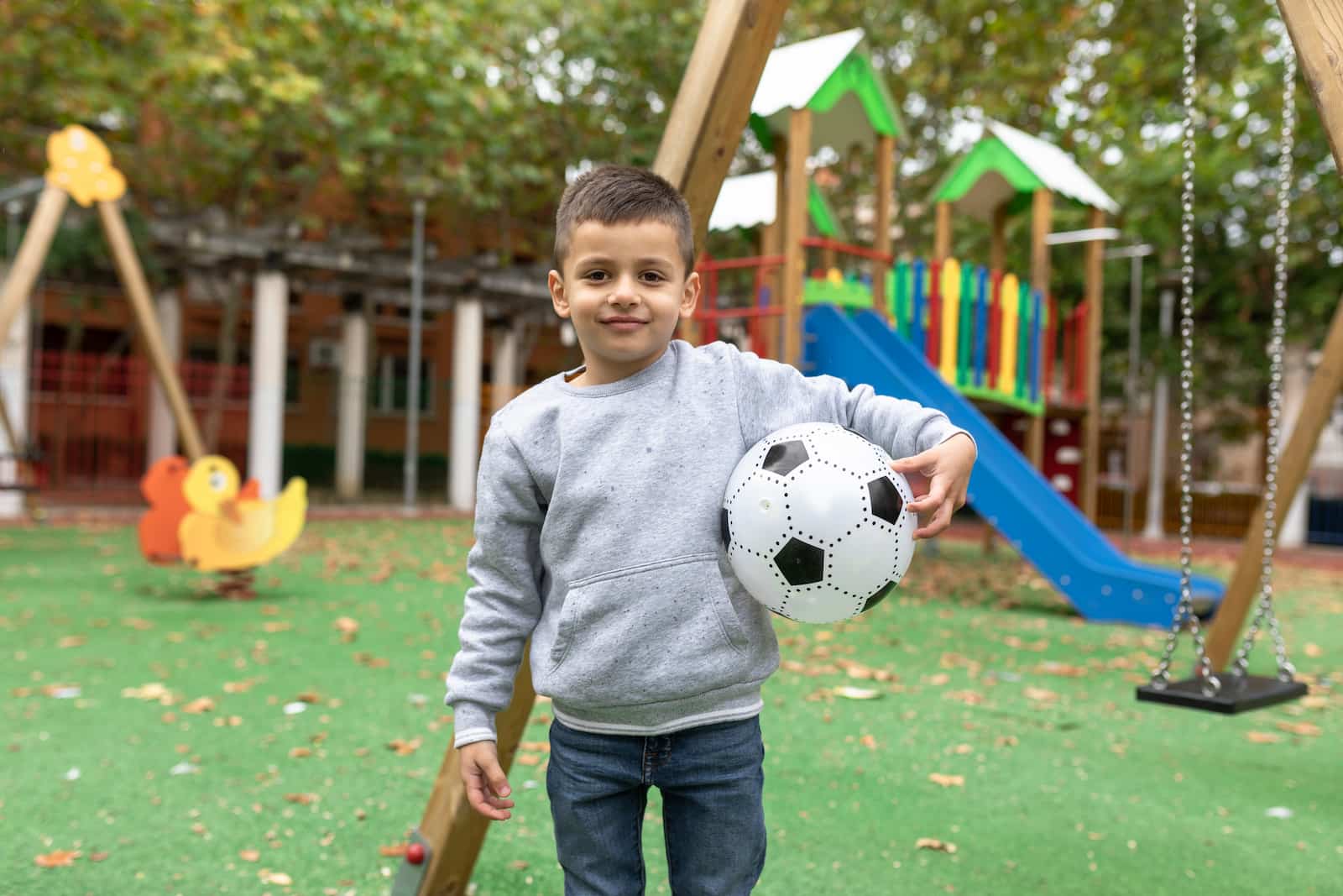 A child standing in a park holding a soccer ball under his arm. 