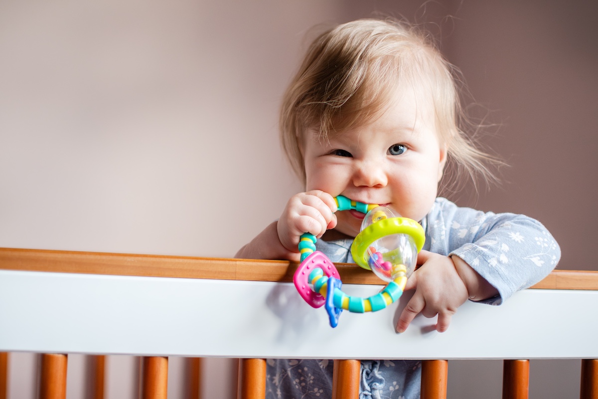 A young baby in her crib biting on a colorful rattle toy. 