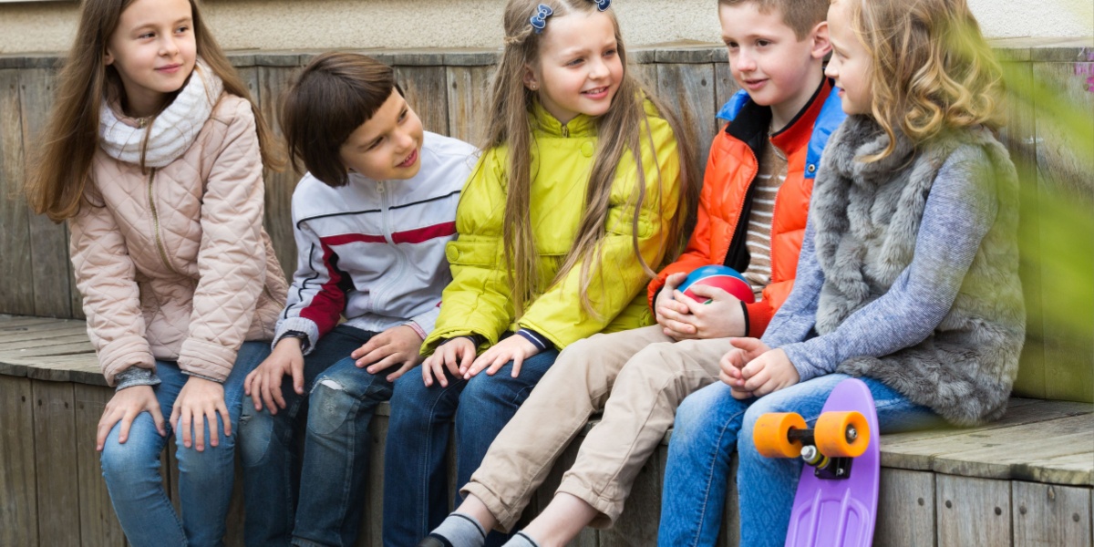 Children sitting and talking in a line in the cool air on a wooden bench. 