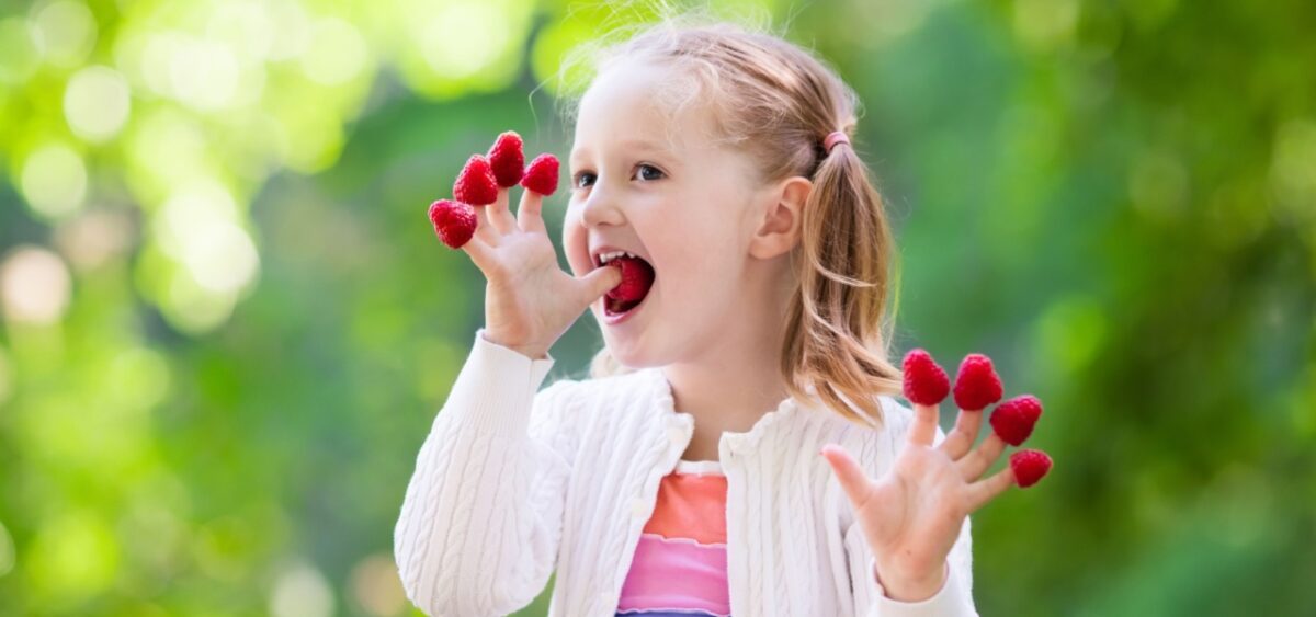 A young child with raspberries on her fingers eating them in a fun sensory activity. 
