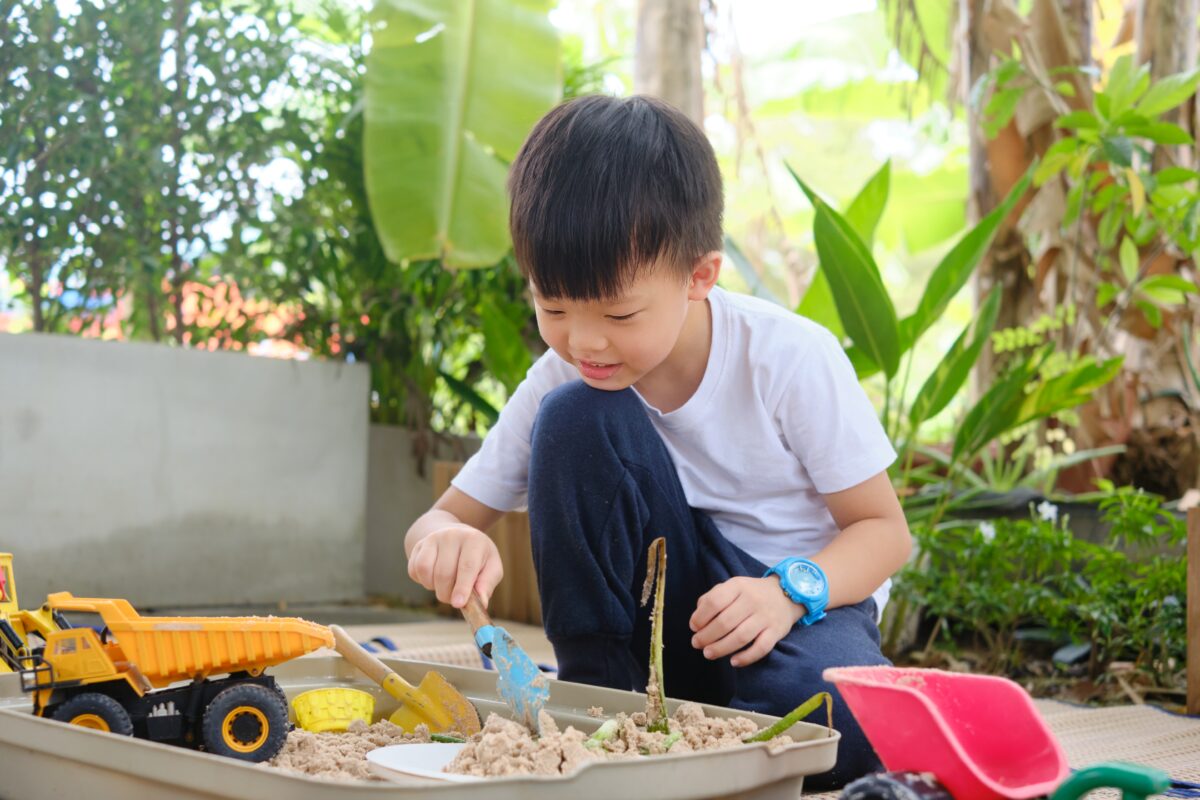 A young boy playing in a box of sand with toy trucks and buckets. 