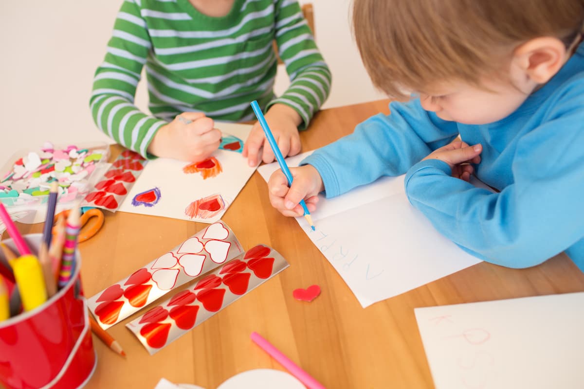 Two young children sitting at a table making hand written cards with heart stickers. 