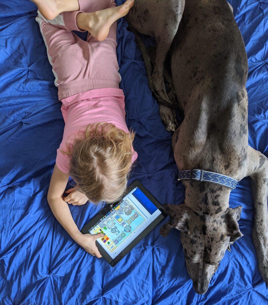 A 3 year old playing alphabet learning games on her i-pad laying on the floor next to her dog. 