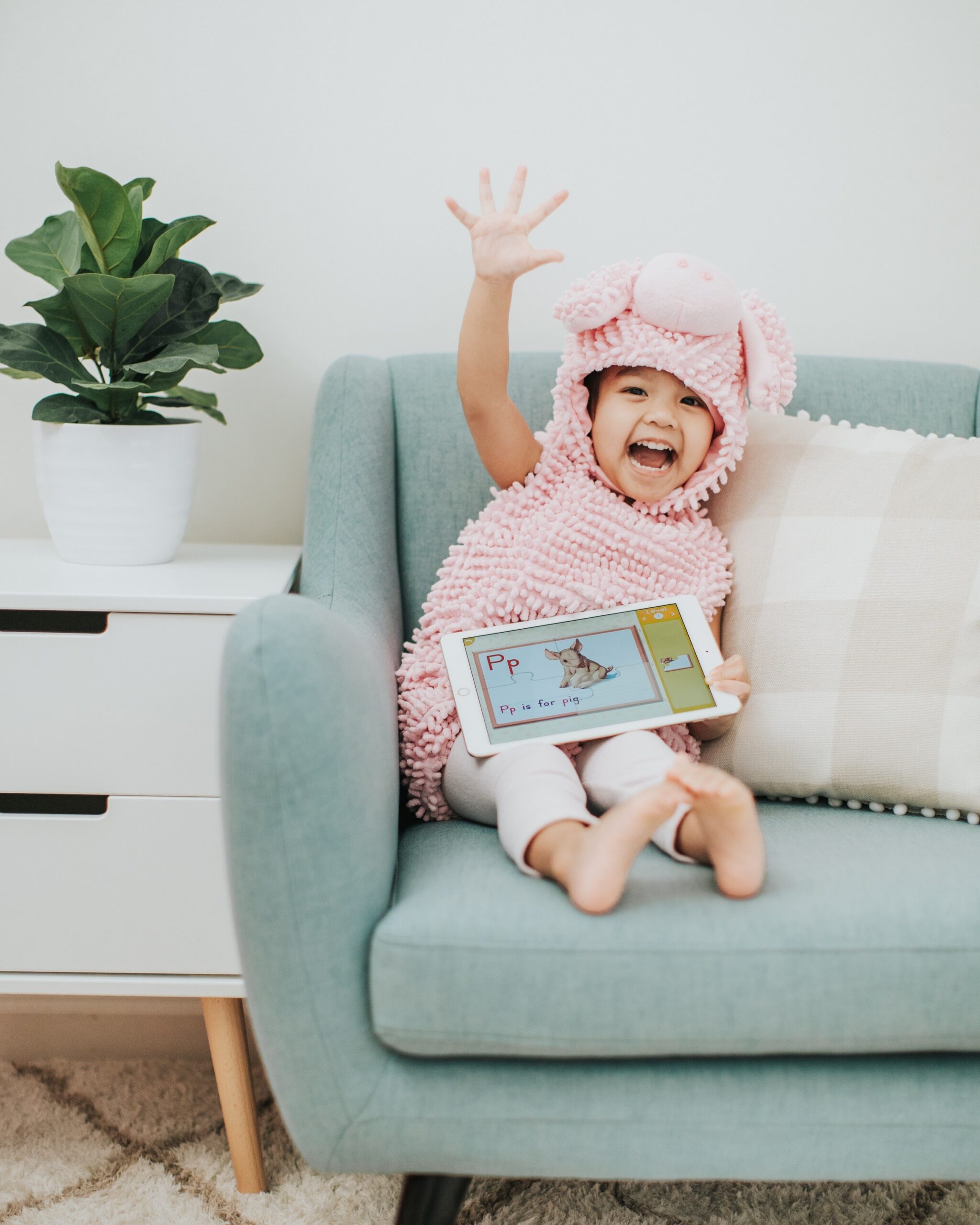 Young girl waving hi while holding an i-pad on her lap that shows her playing the ABCmouse app.