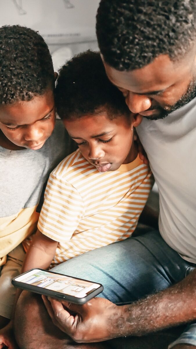 A toddler sitting with their sibling and father looking at the app. 