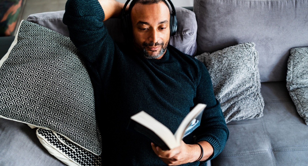 A young father wearing headphones and reading a book while laying on the couch. 