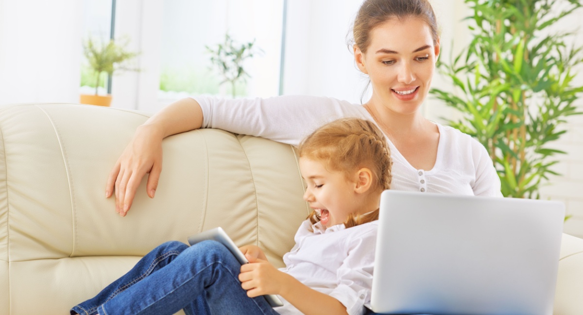 A young girl leaning on her mother while they are both engaged on screens. 