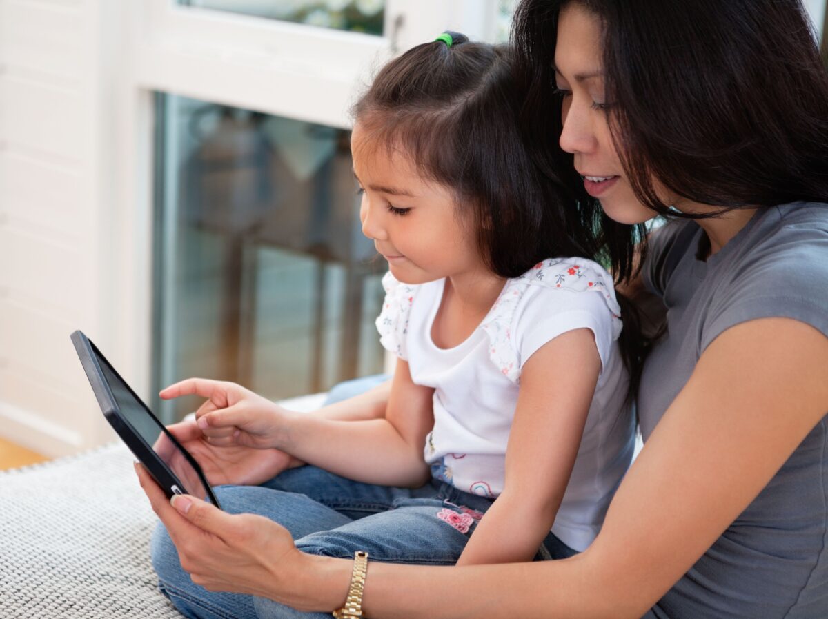 A young girl sitting in her mother's lap holding an i-pad while her mother points to the fun ABCmouse learning game on the screen. 