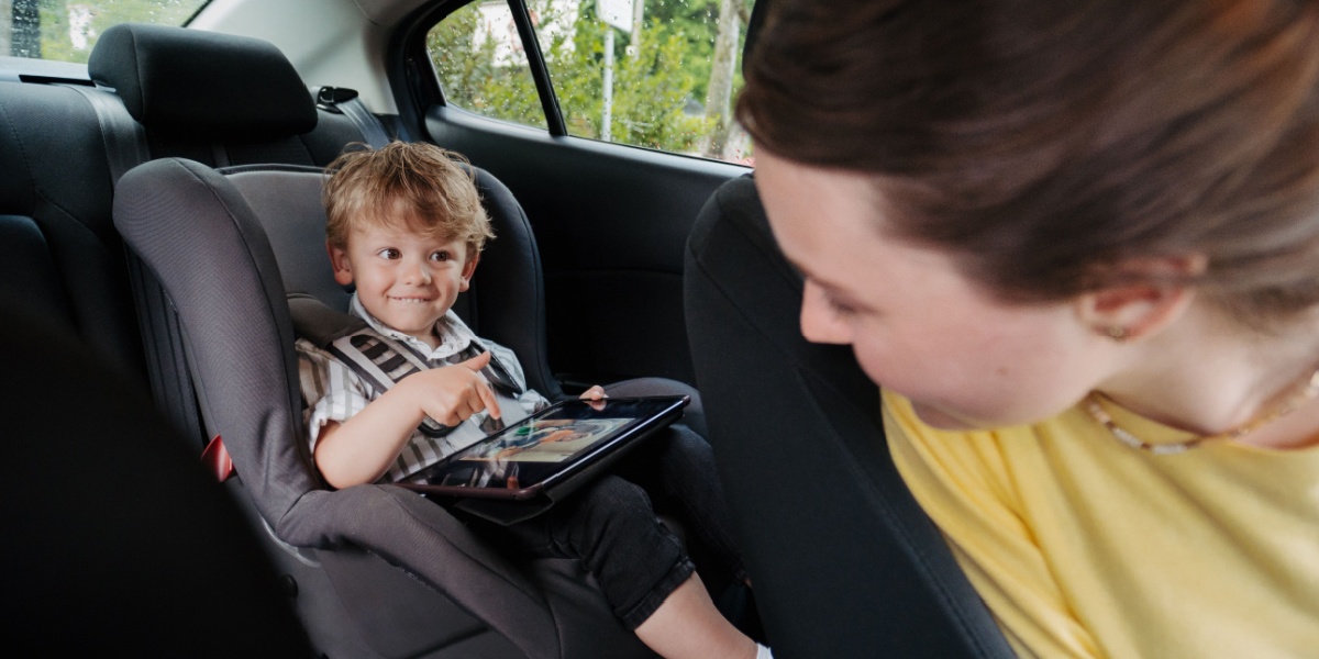 Young boy sitting in his car seat in the back of the car playing ABCmouse app on his i-pad while his mom looks back at him from he drivers seat. 