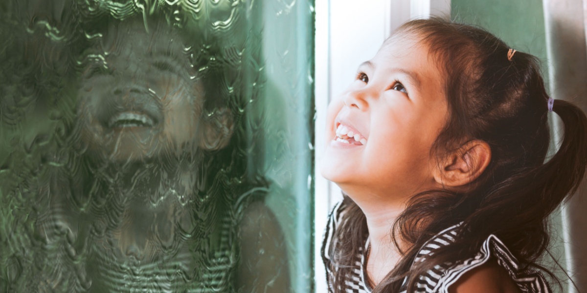 A young girl watching the rain out of a window while smiling.