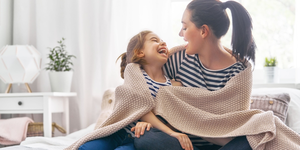 A young mom and daughter wrapped in a blanket together laughing.