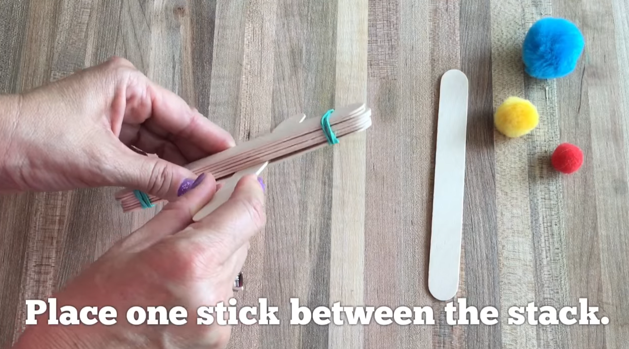 Securing two craft sticks together with a rubber band to create the catapult base