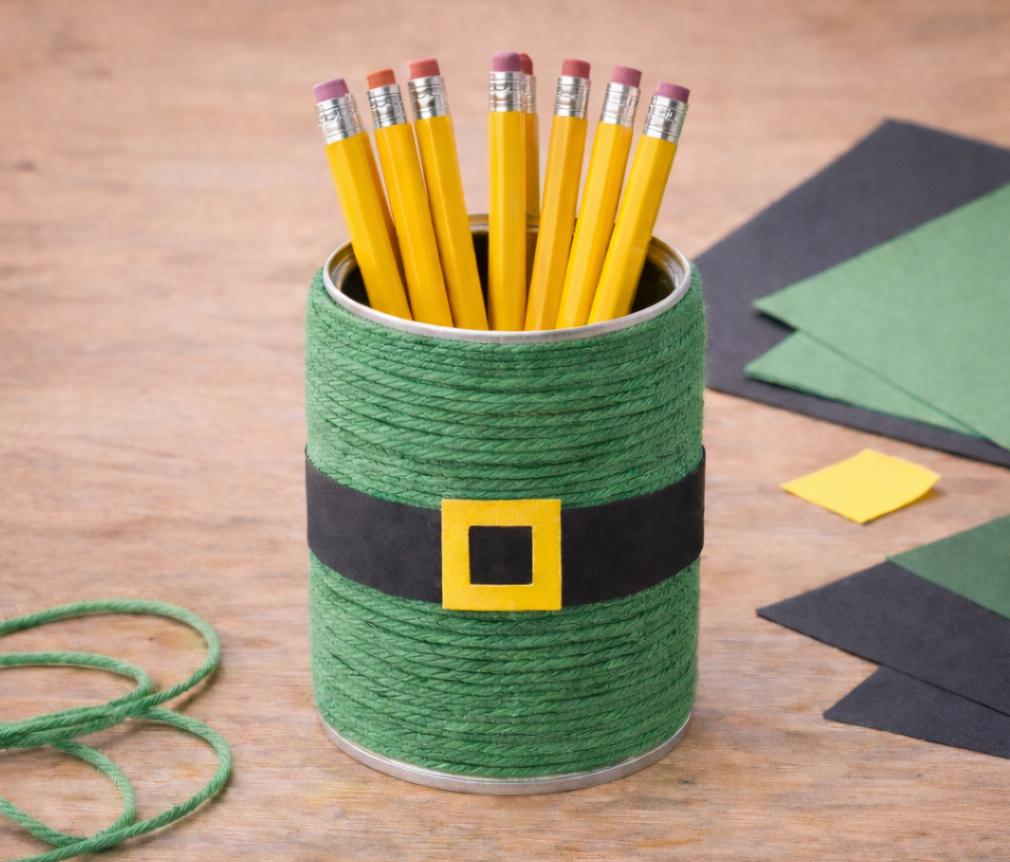 Leprechaun-themed pencil holder wrapped in green yarn with a black belt and yellow buckle, holding yellow pencils on a craft table.