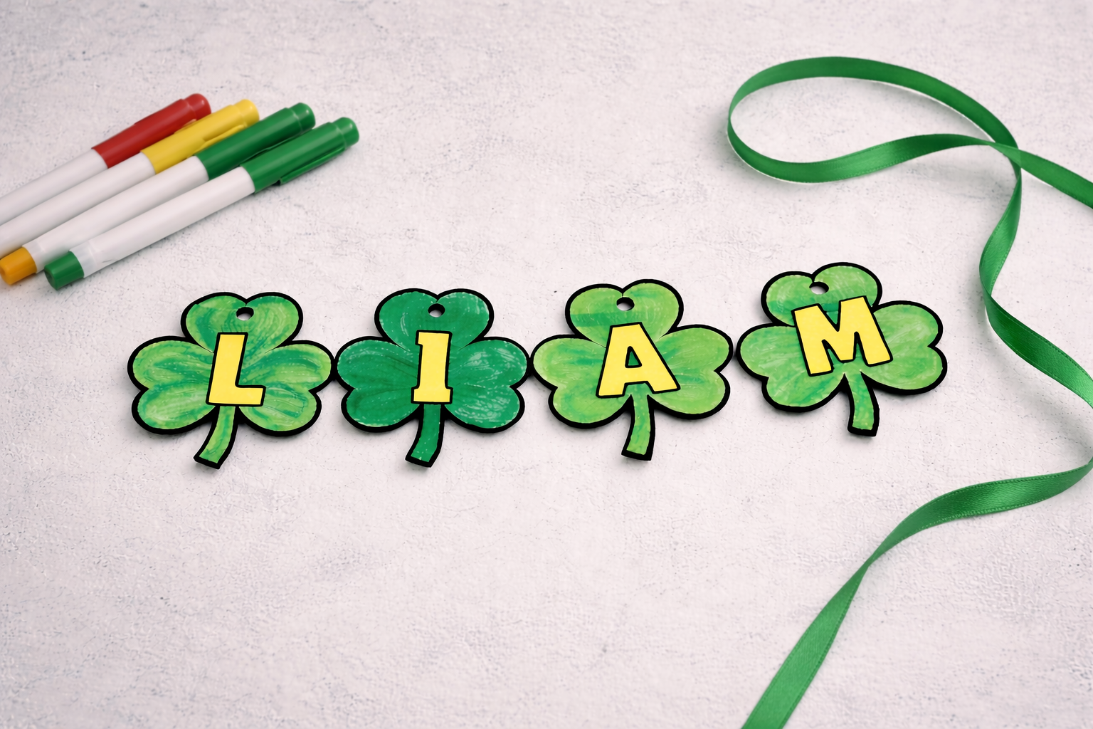 St. Patrick’s Day shamrock name banner spelling “LIAM,” with green shamrock cutouts, ribbon, and markers on a light background.