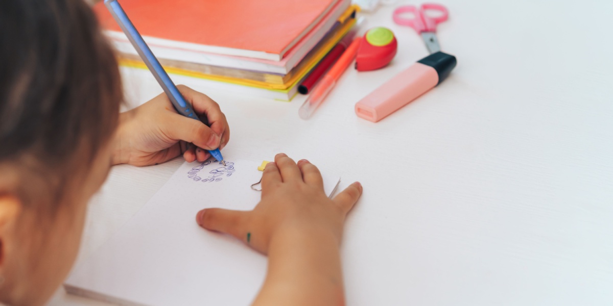 Child drawing on paper with a pencil at a desk with notebooks and school supplies.