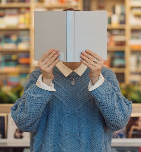 A young child sitting in the library hiding their face behind a light blue book.