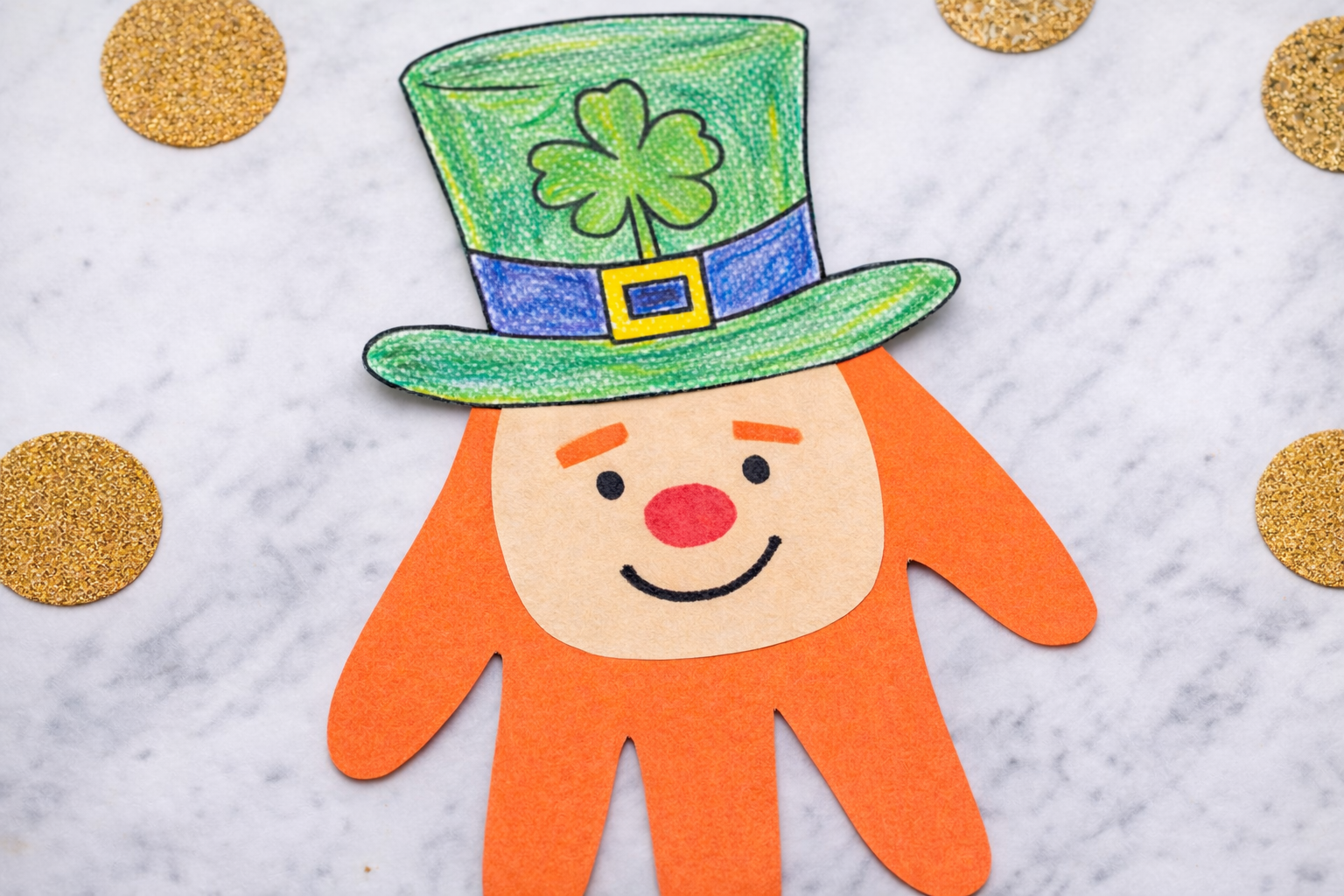 St. Patrick’s Day handprint leprechaun craft with an orange paper hand, smiling face, and a green hat with a shamrock on a marble background.