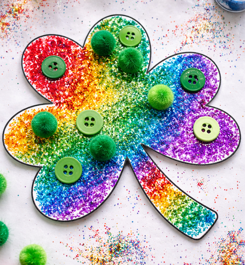 Rainbow shamrock craft covered in glitter with green buttons and pom-poms, scattered with colorful glitter on a white background.