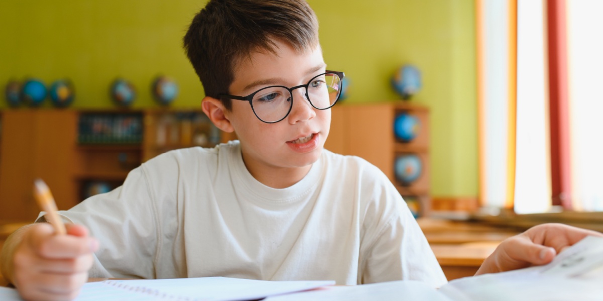 Child wearing glasses reading or working at a desk in a classroom.