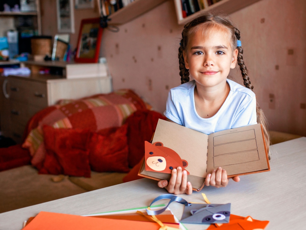 Young girl sitting at a table holding a handmade book and smiling.