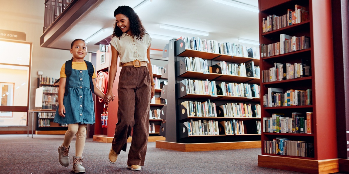 Adult and child walking together through a library holding hands.