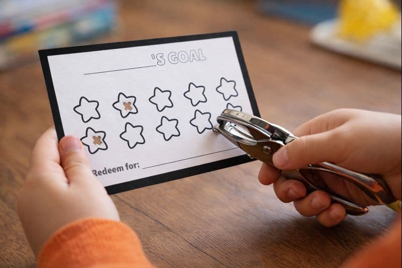 Child’s hands holding a goal reward card while using a hole punch to punch star shapes on the card at a table.