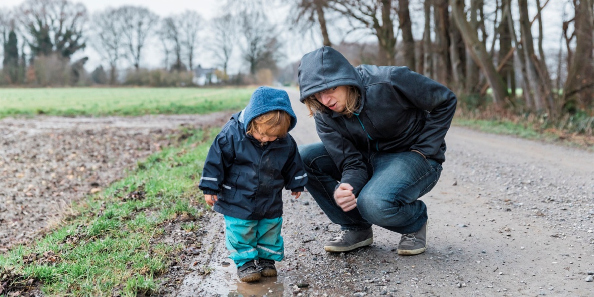 Parent and young child in rain jackets observing puddles along a country path during a spring walk.