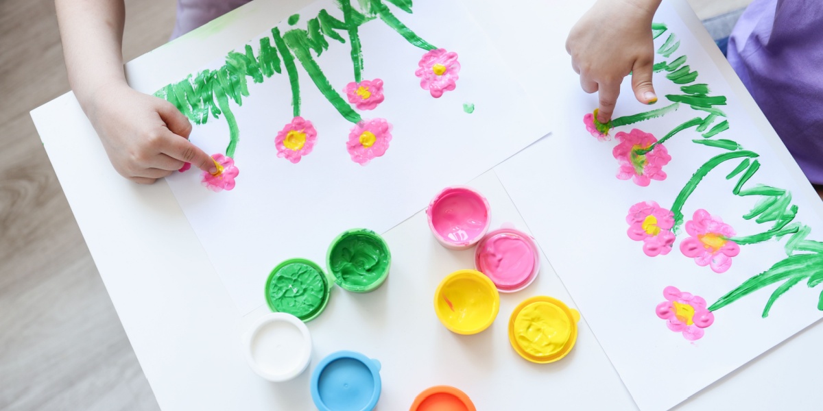 Child using fingerprint painting to create pink flowers with green stems on white paper, with open paint containers nearby.