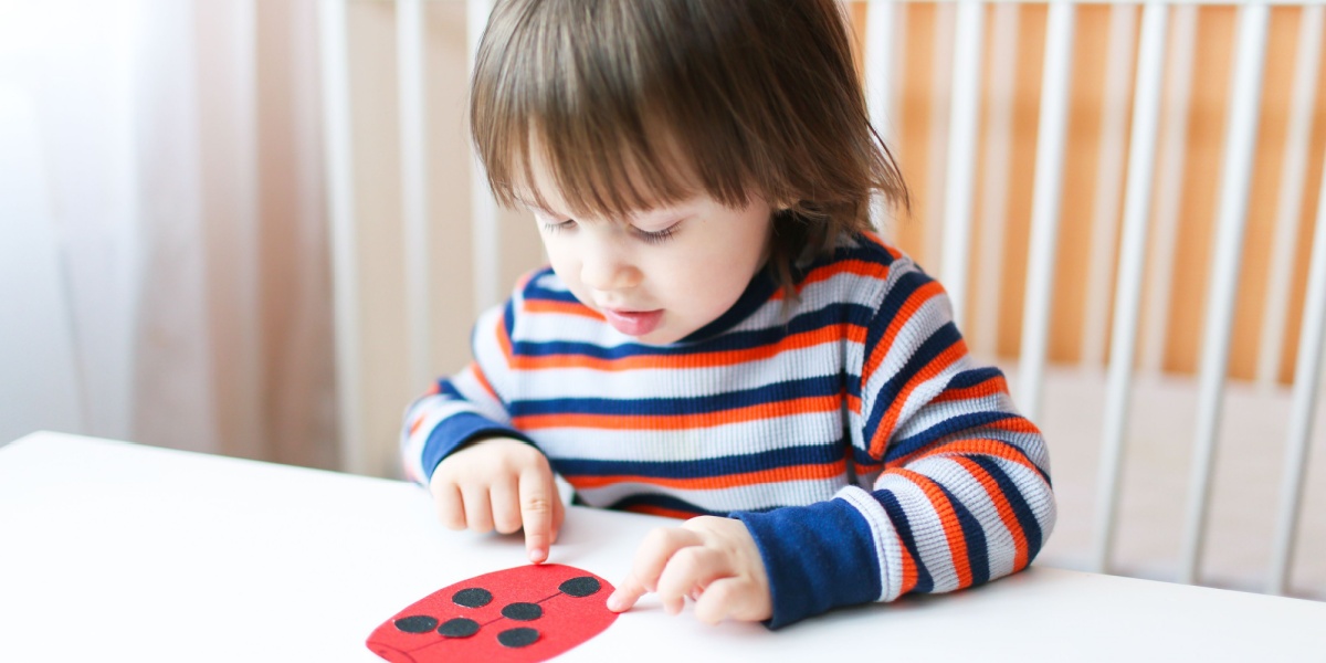 A young child wearing a striped sweater sits at a table, carefully placing black dots onto a red paper ladybug craft.