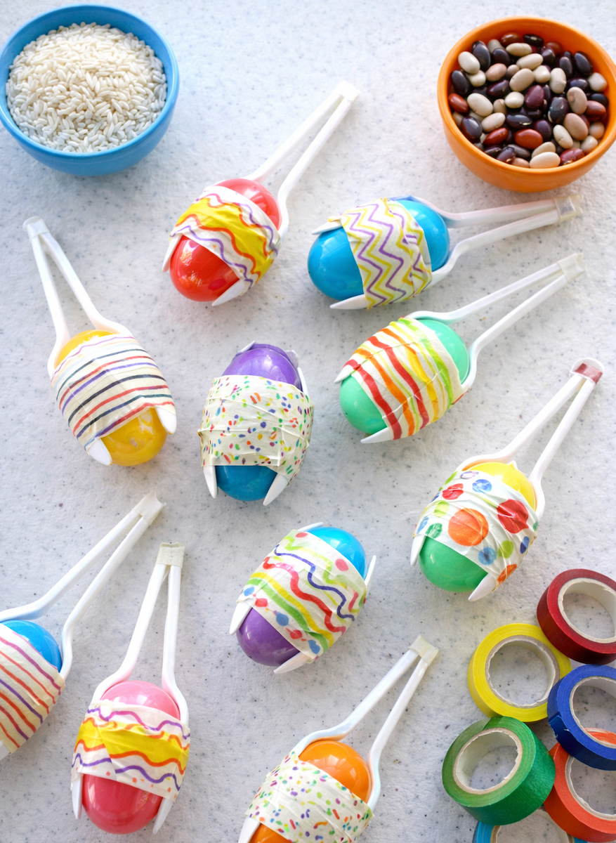 Plastic egg-shaped lollipops decorated with colorful washi tape and drizzle patterns, arranged on a tabletop with craft supplies nearby.