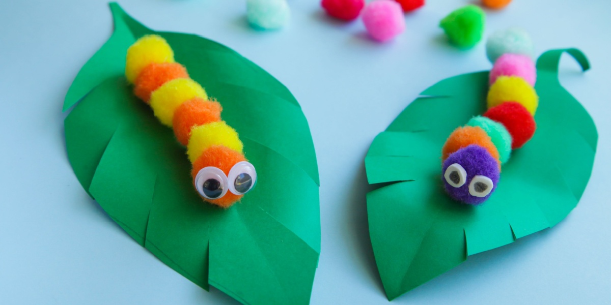Two colorful pom-pom caterpillars with googly eyes resting on large green paper leaves against a light background.