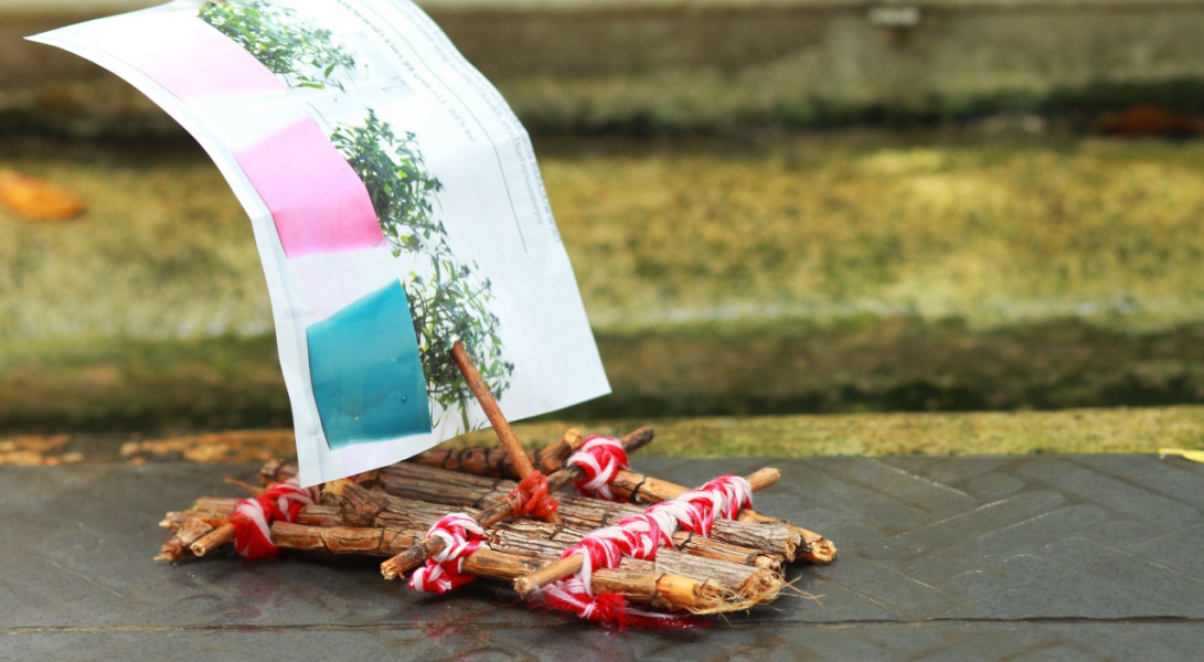 Small handmade raft made of twigs and red string with a paper sail, floating in a shallow stream for a spring outdoor activity.