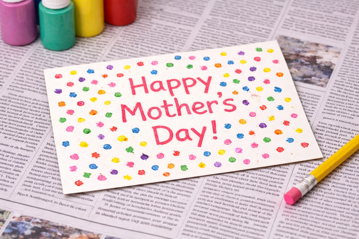 Handmade Mother’s Day card decorated with colorful dot patterns and the message “Happy Mother’s Day!” written in the center, placed on newspaper with craft supplies nearby.