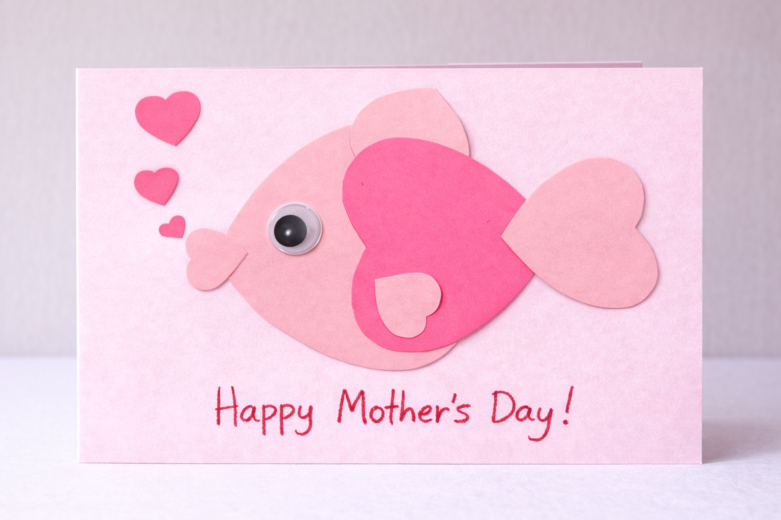 Pink handmade Mother’s Day card featuring a fish made from heart-shaped paper pieces with a googly eye, small hearts floating above it, and the message “Happy Mother’s Day!” written at the bottom.