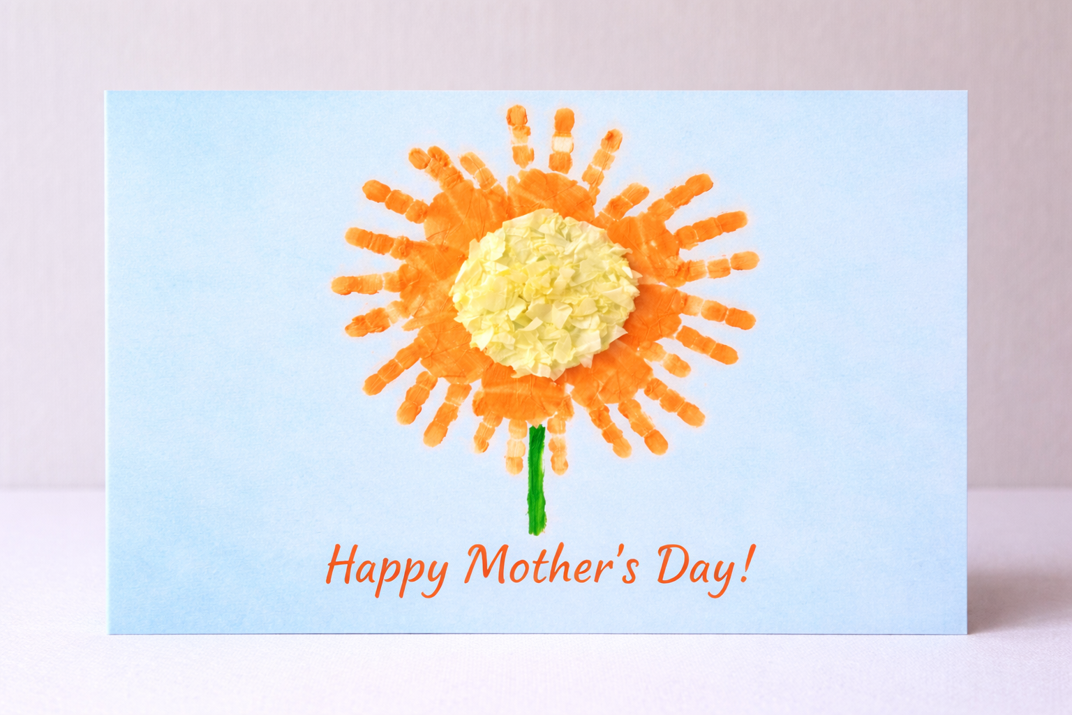 Light blue handmade Mother’s Day card featuring a flower made from orange handprints with a crumpled paper center, a green stem, and the message “Happy Mother’s Day!” written at the bottom.
