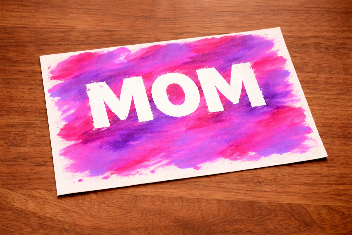 Handmade card with bold white “MOM” lettering over a pink and purple painted background on a wooden surface.