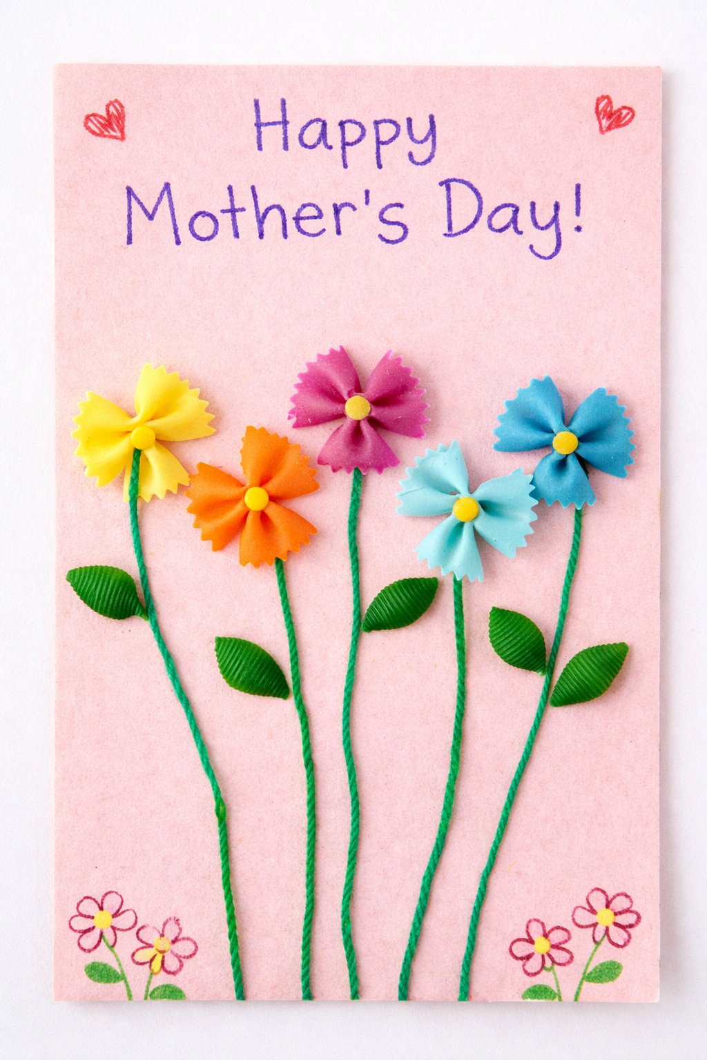 Pink handmade Mother’s Day card featuring colorful flowers made from bowtie pasta, with green stems and leaves, and the message “Happy Mother’s Day!” written at the top with small hearts and flower drawings.