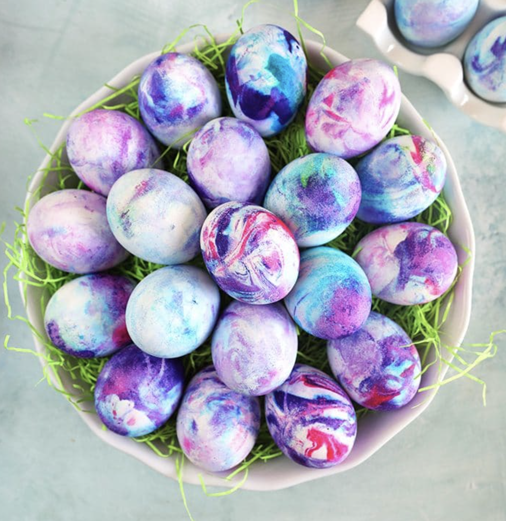 Bowl filled with marbled Easter eggs in shades of purple, blue, and pink, resting on green decorative grass.