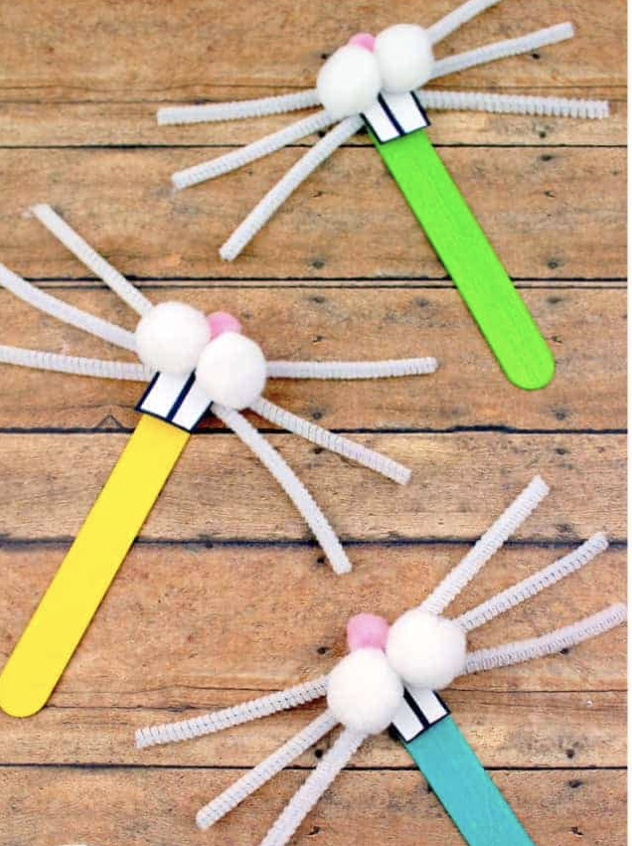 Bunny crafts made from colorful popsicle sticks with white pom-pom faces, pipe cleaner whiskers, and small pink pom-pom noses on a wooden background.